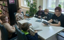 Pięcioro dzieci siedzi przy stole wraz z kobietą, troje z nich czyta książkę. Dw&oacute;jka dzieci patrzy w stronę kobiety.