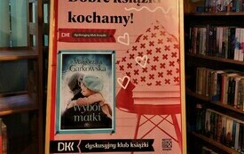 Sztaluga z plakatem Dyskusyjnych Klub&oacute;w Książki, kt&oacute;ry przedstawia okładkę książki, krzesło wraz z książkami oraz poduszką i hasło: &quot;Dobre książki kochamy!&quot;