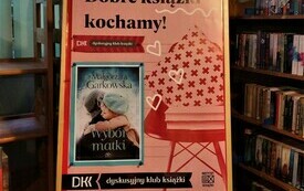 Sztaluga z plakatem Dyskusyjnych Klubów Książki, który przedstawia okładkę książki, krzesło wraz z książkami oraz poduszką i hasło: "Dobre książki kochamy!"