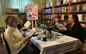 Kobiety siedzą przy stole i trzymają w rękach książki, w tle regały z książkami.