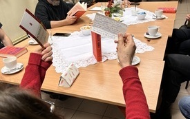 Kilk aosób siedzi przy stole, kobieta trzyma w rękach dwie kartki, a na drugim planie mężczyźni uśmiechają się.