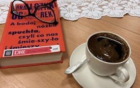 Na stole leży książka, na której leżą okulary, obok filiżanka z kawą.