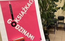 Sztaluga z plakatem Dyskusyjnych Klubów Książki, na sztaludze stoi książka, w tle rośliny.