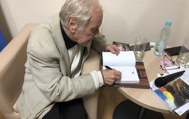 Starszy mężczyzna siedzi w fotelu przy stoliku i podpisuje książkę