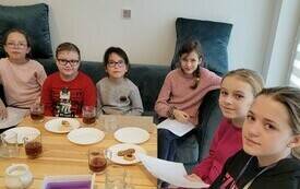 Kilkoro dzieci siedzi przy stole, na którym leżą talerzyki, kubki i czekoladki, uśmiechają się w stronę fotografa.