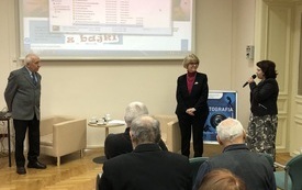 Sala wypełniona uczestnikami spotkania. Na pierwszym planie trzy osoby stoją i przemawiają do uczestników.