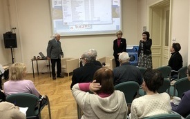 Sala wypełniona uczestnikami spotkania. Na pierwszym planie trzy osoby stoją i przemawiają do uczestników.