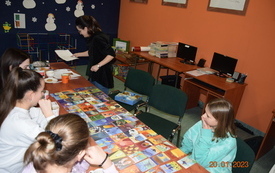 Dziewczynki siedzą przy stole, na kt&oacute;rym rozłożone są karty z r&oacute;żnymi ilustracjami, kobieta trzyma w ręku kartkę papieru.