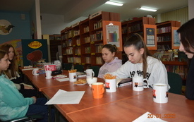 Nastolatki siedzą przy stole, jedna z nich czyta książkę, w tłe są regały książek.