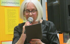 Poetka w okularach trzyma w ręku mikrofon i tekst, czyta. 