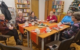 Kilka kobiet siedzi przy stole, na kt&oacute;rym leżą książki, stoją filiżanki i serwetki.