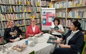 Cztery kobiety siedzą przy stole, jedna z nich trzyma książkę. W tle sztaluga z plakatem Dyskusyjnych Klubów Książki oraz regały wypełnione książkami.