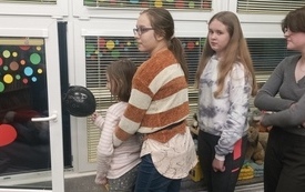 Trzy dziewczynki stoją przy oknie, jedna z nich trzyma w ręce balonik, który wypuszcza przez okno.