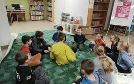 Dzieci siedzą w k&oacute;łku po turecku na dywanie. w środku siedzi chłopiec zwr&oacute;cony plecami do fotografa.