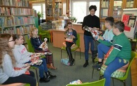 Dzieci siedzą w kole na krzesłach, w rękach trzymają zapakowane upominki, kobieta wręcza prezent jednemu z dzieci