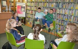 Dzieci siedzą w kole na krzesłach, dwóch chłopców pokazuje okładkę książki.