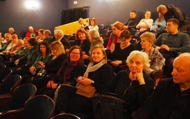 Grupa ludzi siedzi na krzesłach, dwie kobiety uśmiechają się, w tle widownia teatralna, większość miejsc zajętych przez ludzi. 