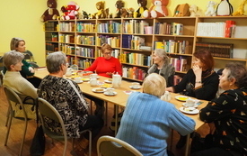 Grupa kobiet siedzi przy stole, cztery z nich są odwr&oacute;cone plecami. W tle regały z książkami, na g&oacute;rnym regale pluszowe zabawki.