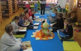 Dzieci siedzą na poduszkach na dywanie, na środku znajduje się pluszowa kaczka. Poza kręgiem dzieci stoi kobieta, która trzyma książkę w ręku.