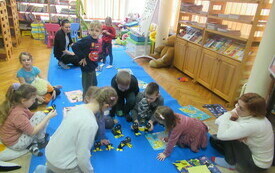Dzieci siedzą na dywanie i trzymają w rękach papierowe ptaki, kt&oacute;re wykonały podczas spotkania. W tle regały z książkami.