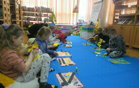 Dzieci siedzą na dywanie i przyklejają do kartek papierowe elementy. 