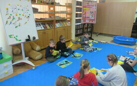 Dzieci siedzą na dywanie i wycinają z papieru r&oacute;żne elementy. W tle tablica magnetyczna, na kt&oacute;rej przypięte są sylwetki ptak&oacute;w. 
