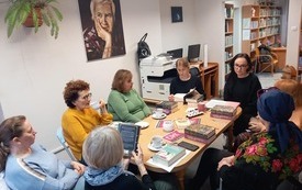Grupa kobiet siedzi dookoła stołu, na kt&oacute;rym leżą książki. W tle portret mężczyzny. 