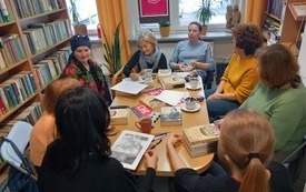 Grupa kobiet siedzi dookoła stołu, rozmawiają, na stole znajdują się książki. 