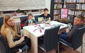 Tr&oacute;jka dzieci i kobieta siedzą przy stole, trzymają w rękach książki, pokazując ich okładki.