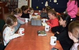 Dzieci siedzą przy stole, na kt&oacute;rym znajdują się książki i kubki z logo Dyskusyjnych Klub&oacute;w Książki, w tle dekoracja zimowa z bałwankami. 