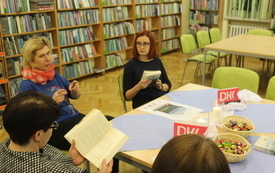 Trzy kobiety siedzą przy stole, jedna ma otwartą książkę, a kolejna żywo gestykuluje, opowiadając.