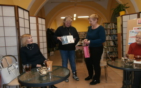 Dwie kobiety siedzą przy stolikach, za nimi stoją dwie kobiety jedna z nich trzyma prezent, a druga książki