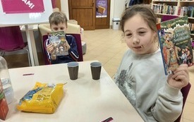 Dzieci siedzą przy stoliku i pokazują książkę do osoby robiącej zdjęcie.
