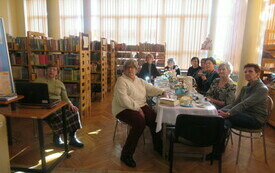 Grupa senior&oacute;w siedząca przy stole, na drugim planie regały z książkami.