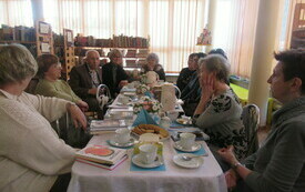 Grupa senior&oacute;w siedząca przy stole, na drugim planie regały z książkami.