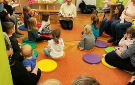 Dzieci siedzą na dywanie, patrzą na panią, kt&oacute;ra czyta i pokazuje bajkę. 