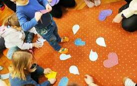 Dzieci w k&oacute;łku siedzą na dywanie i rozkładają serduszka, obok pani prowadząca spotkanie.