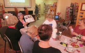 Kobiety siedzą przy stole, jedna z nich pokazuje książkę i opowiada, w tle ekran z prezentacją multimedialną.