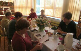 Grupa kobiet siedzi przy stole, jedna z nich pochylona trzyma w ręku kartkę. W tle okna z żaluzjami i regał z książkami. 