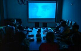 Kobiety siedzą przy stole i oglądają film na dużym ekranie. W sali jest ciemno.