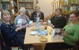 Pięć kobiet siedzi przy stole, dwie z nich przegląda książki. W tle regały z książkami. 