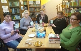 Pięć kobiet siedzi przy stole, troje z nich trzyma w ręku książkę, w tle regały z książkami. 