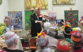 Goście spotkania w kolorowych wiankach na głowach obserwują starszego mężczyznę grającego na skrzypcach. W tle obrazy na sztalugach.