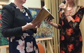 Dwie stojące kobiety w wiankach na głowach i kwiecistych sukienkach trzymają w rękach książki. W tle obrazy na sztalugach.
