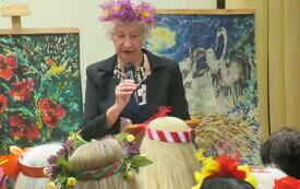 Kobieta w starszym wieku w koralach i wianku na głowie trzyma mikrofon. W tle obrazy na sztalugach.