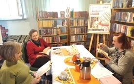 Trzy kobiety siedzą przy stole, trzymają w rękach otwarte książki. W tle regały z książkami. 