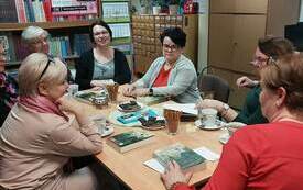 Grupa kobiet siedzi przy stole, na kt&oacute;rym leżą książki, ciastka i filiżanki. W tle plakat Dyskusyjnych Klub&oacute;w Książki, kalendarz ścienny oraz regały z książkami.  