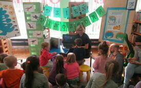 Dzieci biorą udział w spotkaniu na temat dinozaur&oacute;w. Bibliotekarka trzyma w ręku komputer.  W tle prace przedstawiające dinozaury. 