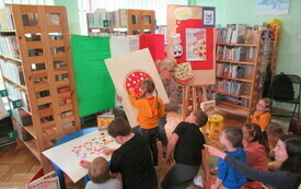 Dzieci biorą udział w spotkaniu na temat dnia pizzy. Bibliotekarka prezentuje obraz pizzy. W tle flaga Włoch. 