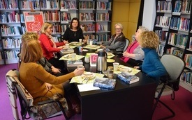 Przy stole kobiety siedzą i rozmawiają, na stole książki, za plecami moderatorki tablica z informacją o spotkaniu. 